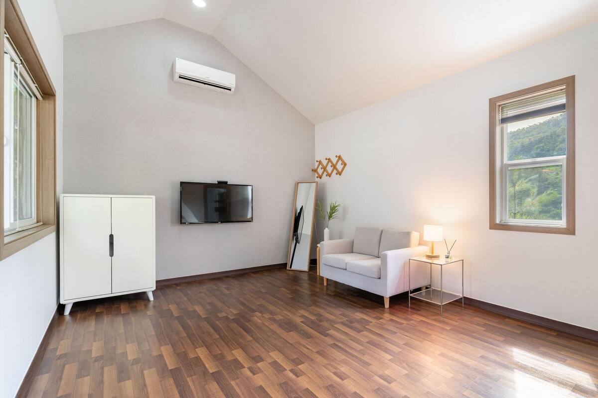 Una habitación amplia con suelo de madera, un sofá blanco, un televisor fijado a la pared y una ventana con vistas a árboles.