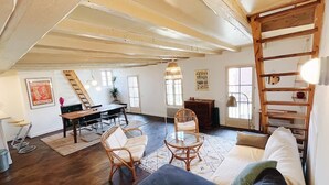 A spacious loft with a high ceiling, exposed beams, a dining area with a table and chairs, a sofa, and a ladder shelf. - Loft in heart of Annecy with balcony over river (Annecy)