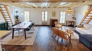 A spacious loft with a high ceiling, exposed wooden beams, a dining area with a long table and chairs, a seating area with wicker furniture, and large windows allowing natural light. - Loft in heart of Annecy with balcony over river (Annecy)