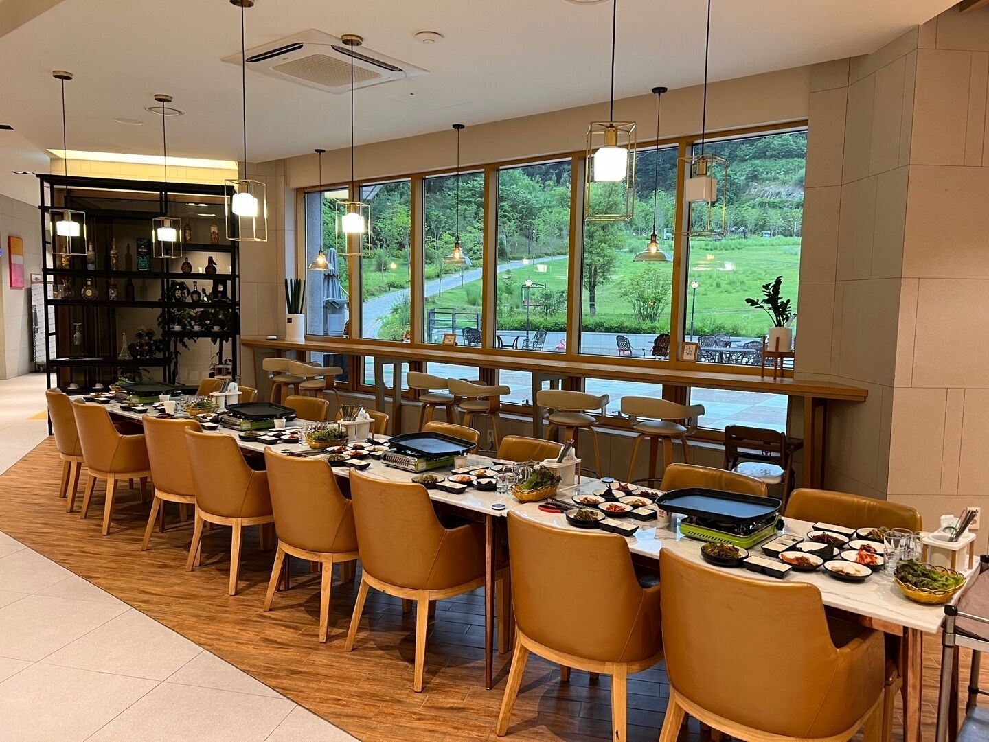 A dining area with multiple tables and chairs, large windows with a view of greenery, and pendant lighting.