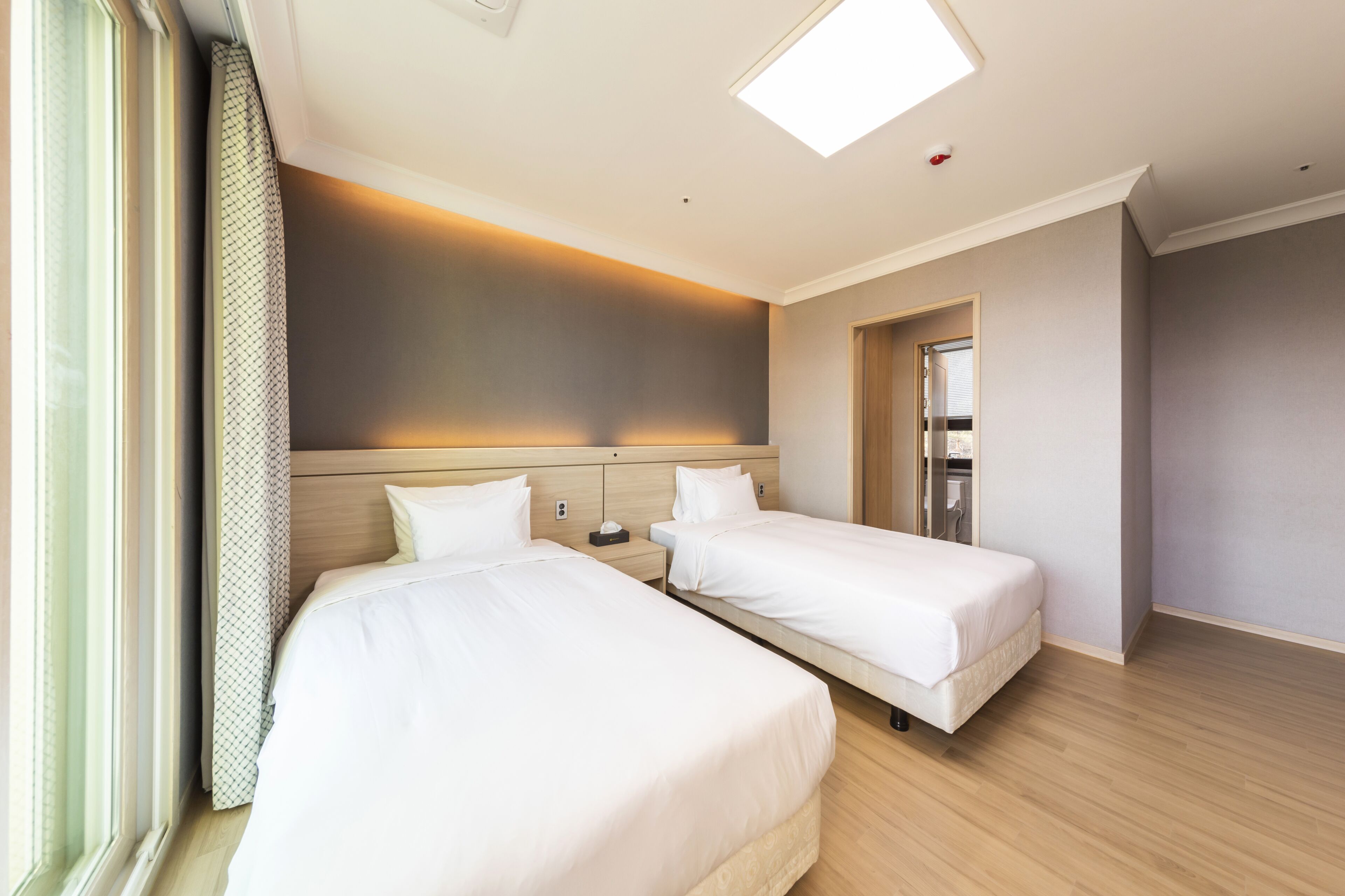 Habitación de hotel con dos camas, suelo de madera, un ventanal grande con cortinas y una lámpara en el techo.