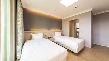 Habitación de hotel con dos camas, suelo de madera, un ventanal grande con cortinas y una lámpara en el techo.
