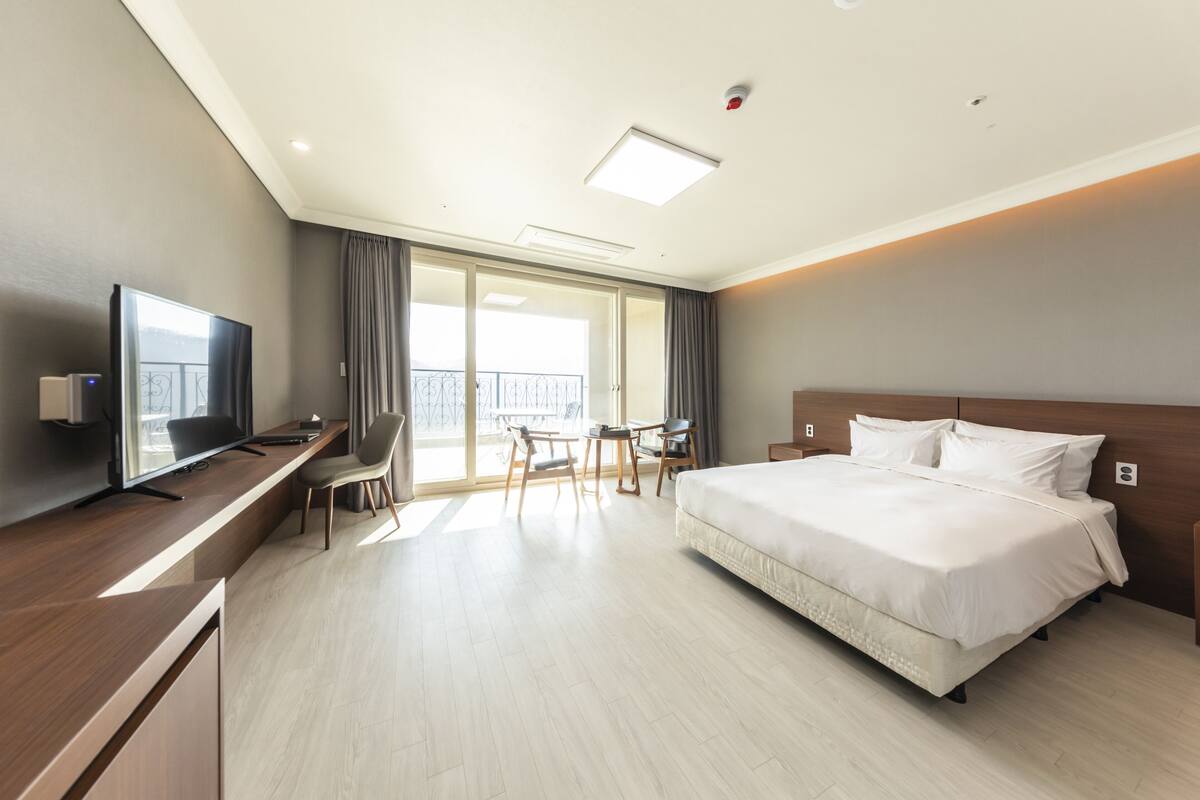 A modern hotel room with a large bed, a desk with a TV, a balcony with a view, and a sliding door.