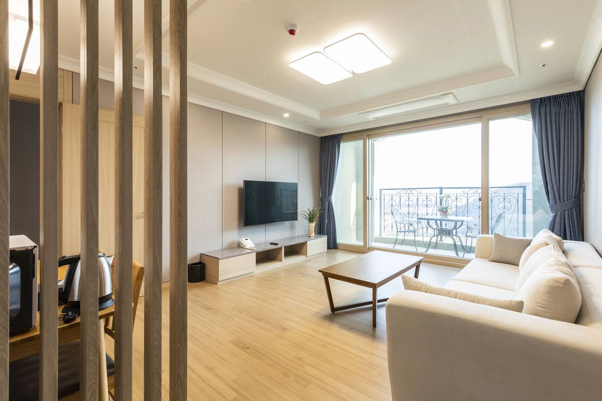 A modern living room with a sofa, a coffee table, a TV, and a balcony with a view.
