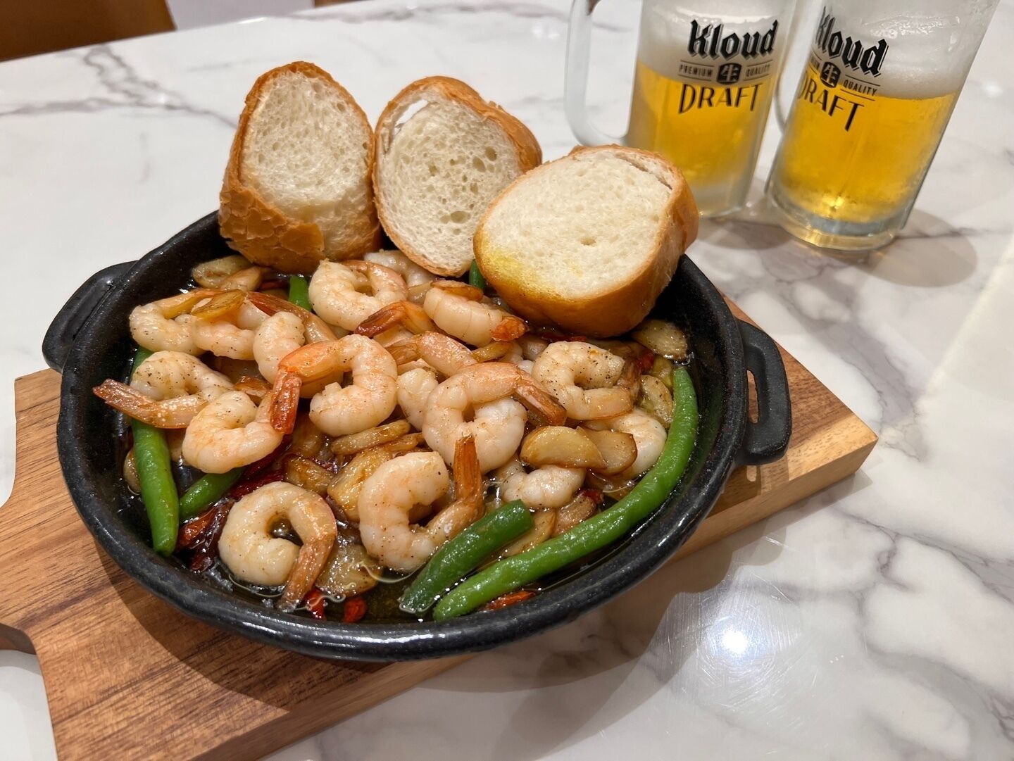 A skillet with cooked shrimp and green beans, accompanied by two slices of bread and two glasses of beer.