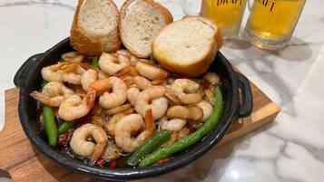 A skillet with cooked shrimp and green beans, accompanied by two slices of bread and two glasses of beer.