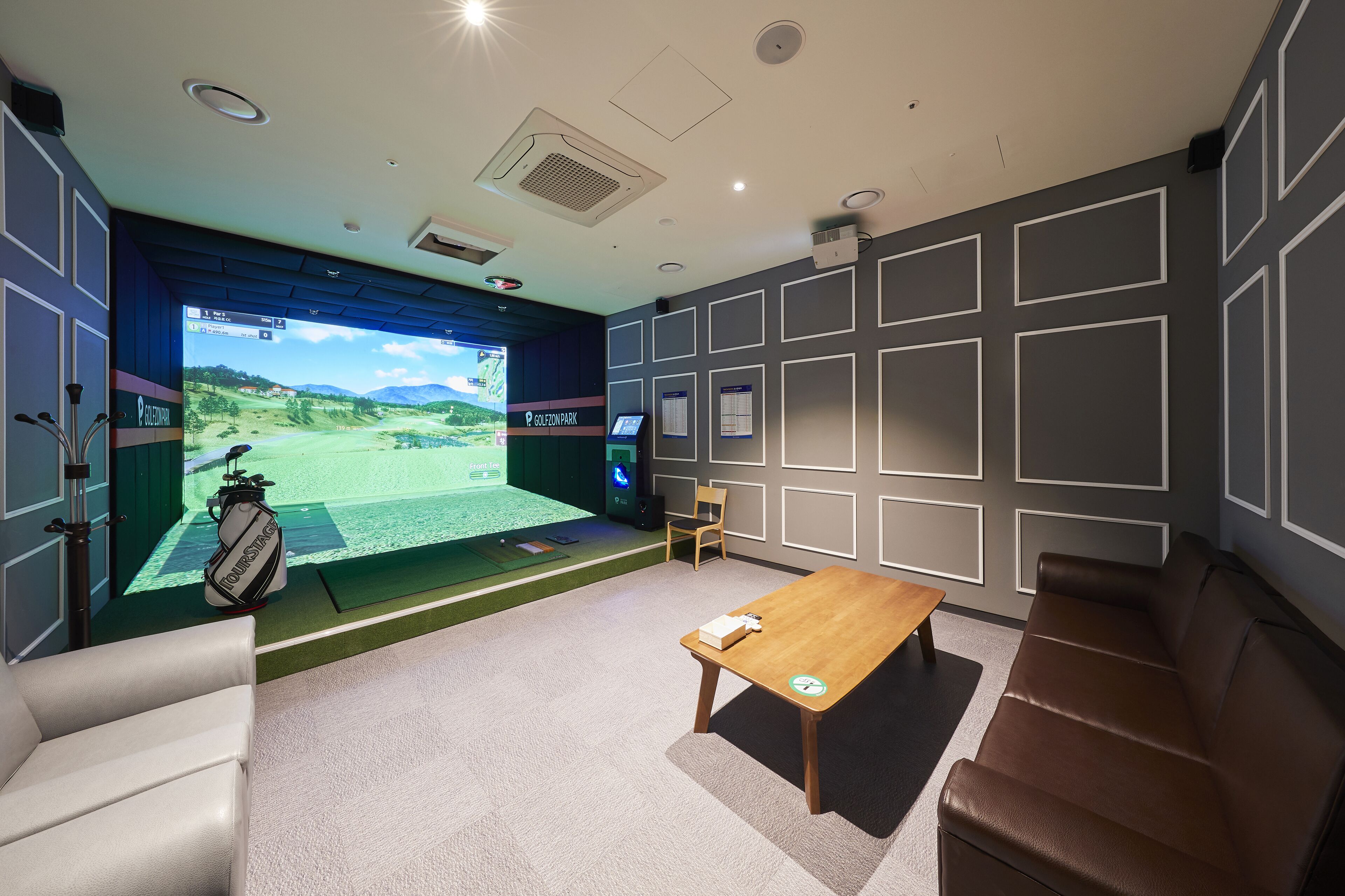 Una habitación con equipo de simulación de golf, una mesa de madera, un sofá marrón y una silla.