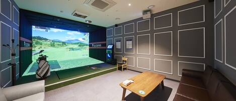 Una habitación con equipo de simulación de golf, una mesa de madera, un sofá marrón y una silla.