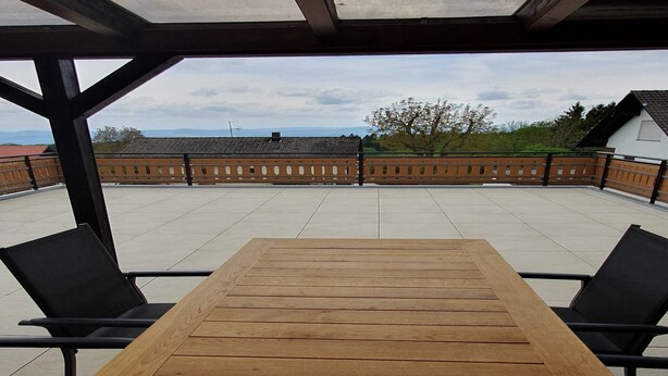 En overdækket terrasse med et træbord og sorte stole, der giver udsigt til bygninger og træer.