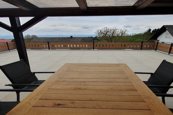 A covered patio with a wooden table and black chairs, offering a view of buildings and trees. - Elegante Ferienwohnung im Schwarzwald, Görwihl (Görwihl)