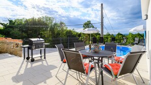 Une terrasse extérieure équipée d’un barbecue, d’une table à manger, de chaises et d’une piscine en arrière-plan.