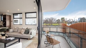 A modern living room with a sofa, coffee table, and chairs, a balcony with a railing, and a city view. - Pet Friendly - Sport Precinct Luxury Style over four levels (Cremorne)