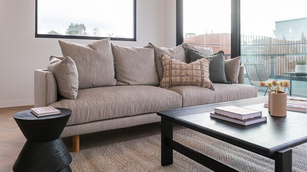 A modern living room with a beige sofa, a black coffee table, and a view of a cityscape through large windows. - Pet Friendly - Sport Precinct Luxury Style over four levels (Cremorne)