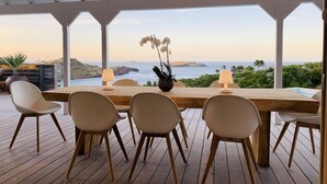 Una terraza de madera con una mesa y sillas, con vista a un paisaje costero con colinas y el océano.