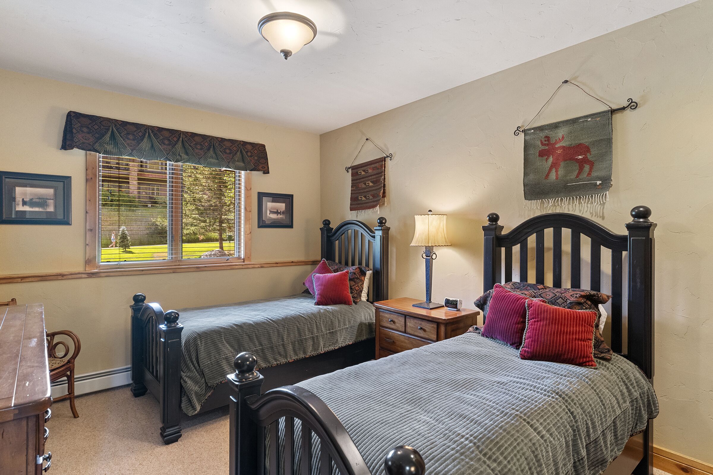 A bedroom with two single beds, a wooden dresser, a nightstand, a lamp, and a window with blinds.