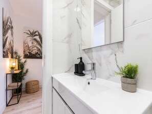 A modern bathroom with a white marble sink, a black soap dispenser, and a large mirror. There are two potted plants on the counter and a woven basket on the floor.