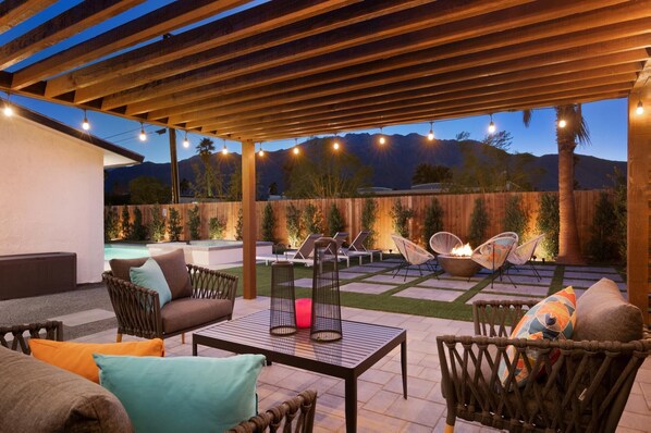 Une terrasse couverte, aménagée avec du mobilier d’extérieur, un foyer et offrant une vue sur une chaîne de montagnes au crépuscule.