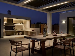 An outdoor dining area with a large table and chairs, a built-in grill, and a modern kitchen with a hanging shelf. - Anthis Luxury Villa (Zakynthos)