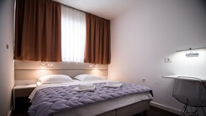 A neatly made bed with a headboard, a bedside table with a lamp, a chair, and a window with curtains.