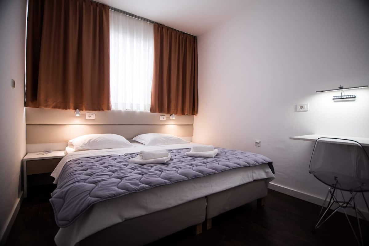 A bedroom with a bed, a desk, a chair, a window with curtains, and a wall-mounted light.