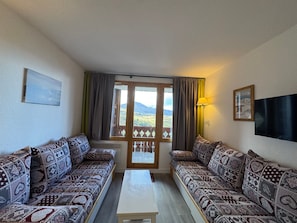 A living room with patterned sofas, a coffee table, a TV, and a balcony with curtains.