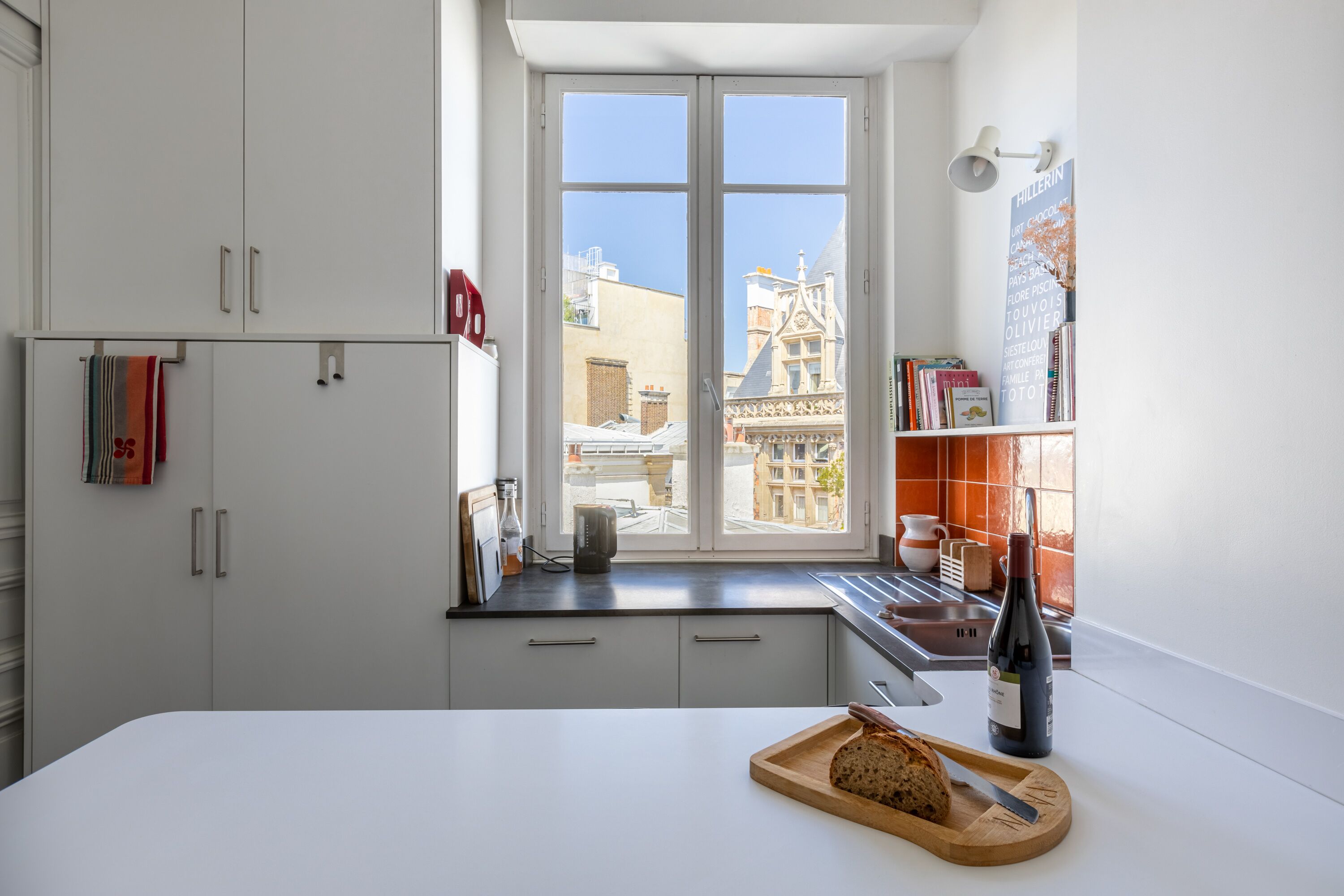 A modern kitchen with a large window, white cabinets, and a wooden cutting board with bread.