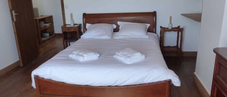 Una cama bien hecha con sábanas blancas y dos toallas, cabecero de madera, dos mesitas de noche con lámparas y un armario de madera.