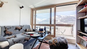 A living room with a sofa featuring animal prints, a dining table with plates and cutlery, a fluffy dog, and a large window with a mountain view. - Modern 3-room apartment, ski-in/ski-out, balcony, exceptional view (Les Menuires)