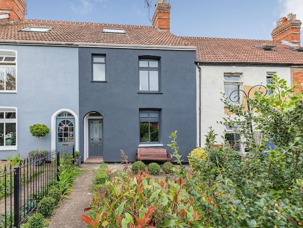 A two-story house with a grey facade, a front door, and a small garden with various plants and a bench.