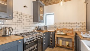 A compact kitchen with a gas stove, oven, and a microwave. There is a wooden table with a tray and a kettle. The kitchen has a tiled backsplash and a stone floor. - 7 Severn Terrace (Watchet)