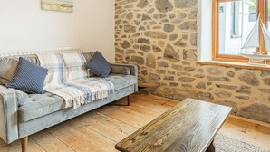 A cozy living room with a stone wall, a wooden coffee table, a grey sofa with patterned cushions, and a window with a sailboat model. - 7 Severn Terrace (Watchet)
