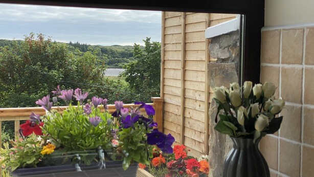 A balcony with a view of a lake and hills, a potted plant with purple flowers, and a vase with white flowers.