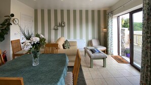 A dining area with a table set for two, a sofa, and a chair. There is a plant on the table and a view of the outdoors through a glass door.