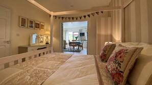 A bedroom with a large bed, a bedside table, a mirror, and a hanging light fixture. There is a door leading to a dining area with a table and chairs.