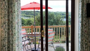 A balcony with a table and chairs, a red umbrella, and a view of a forested area.