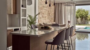 A modern kitchen with a central island, bar stools, pendant lighting, and a stone accent wall. - Anthis Luxury Villas - 3 Bedroom Villas with Private Pool (Zakynthos)
