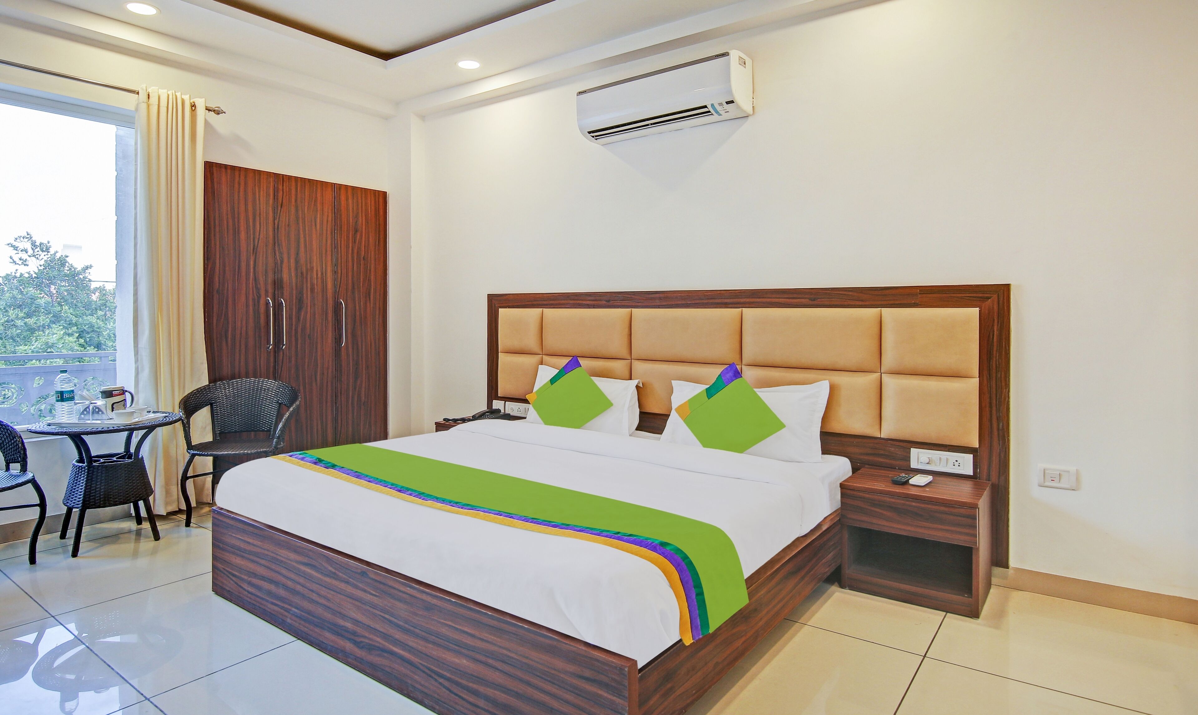 A hotel room with a bed, a wooden wardrobe, a small table with chairs, and an air conditioning unit.