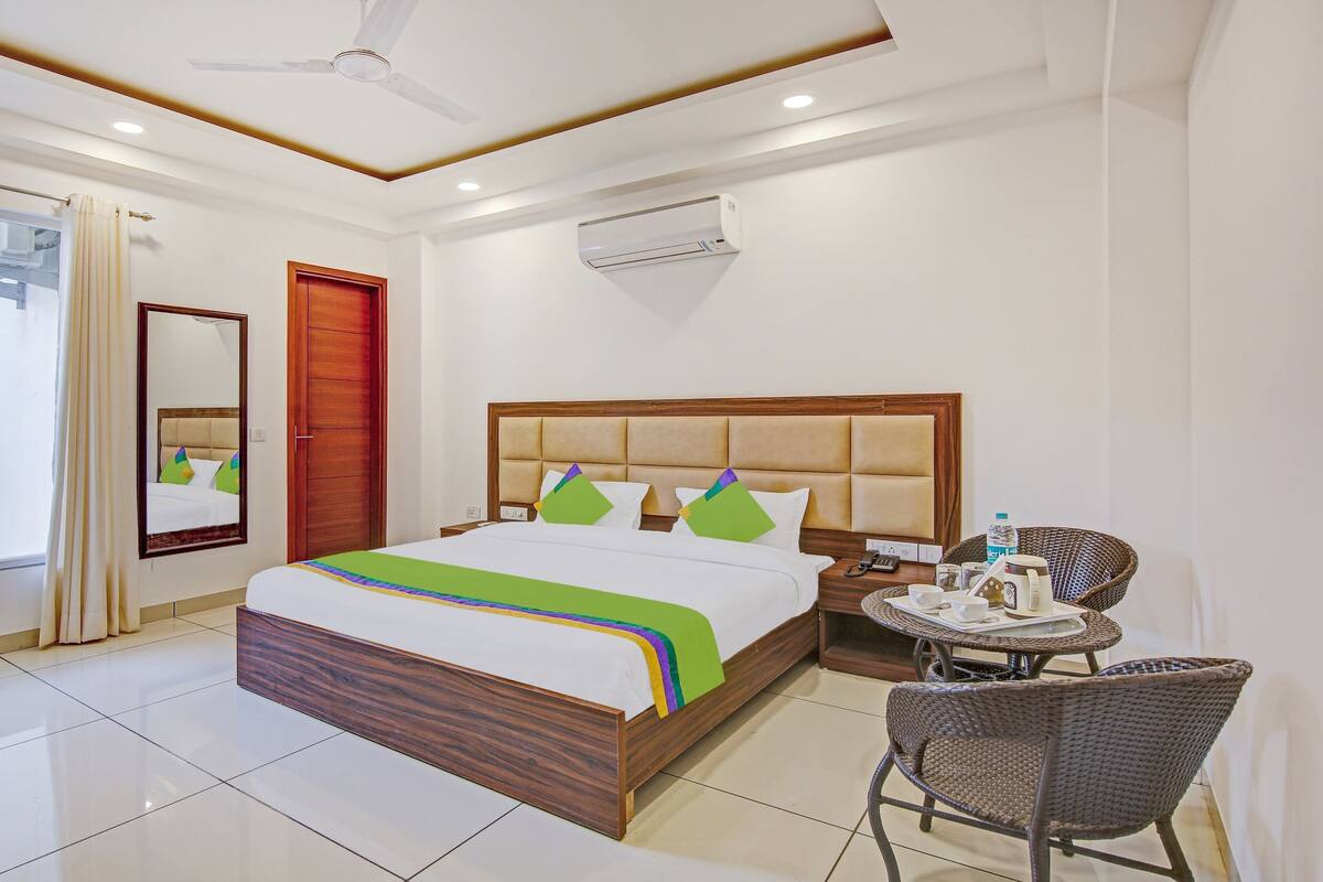 Quarto de hotel com uma cama grande, cabeceira de madeira, mesa de cabeceira com telefone e cesta, cadeira de vime e uma janela com cortinas.