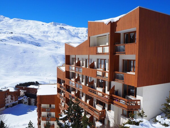 A modern multi-story hotel with wooden balconies and large windows, set against a snowy mountain backdrop.
