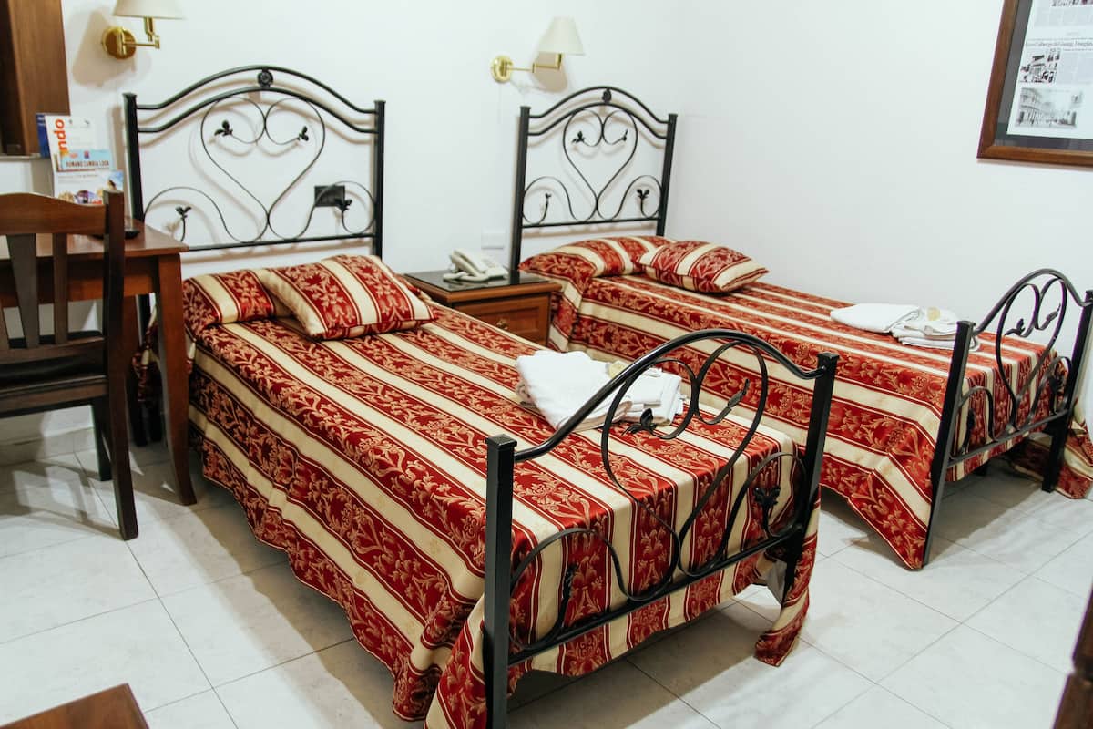 A hotel room with two beds, a wooden chair, a nightstand, and a wall-mounted lamp.