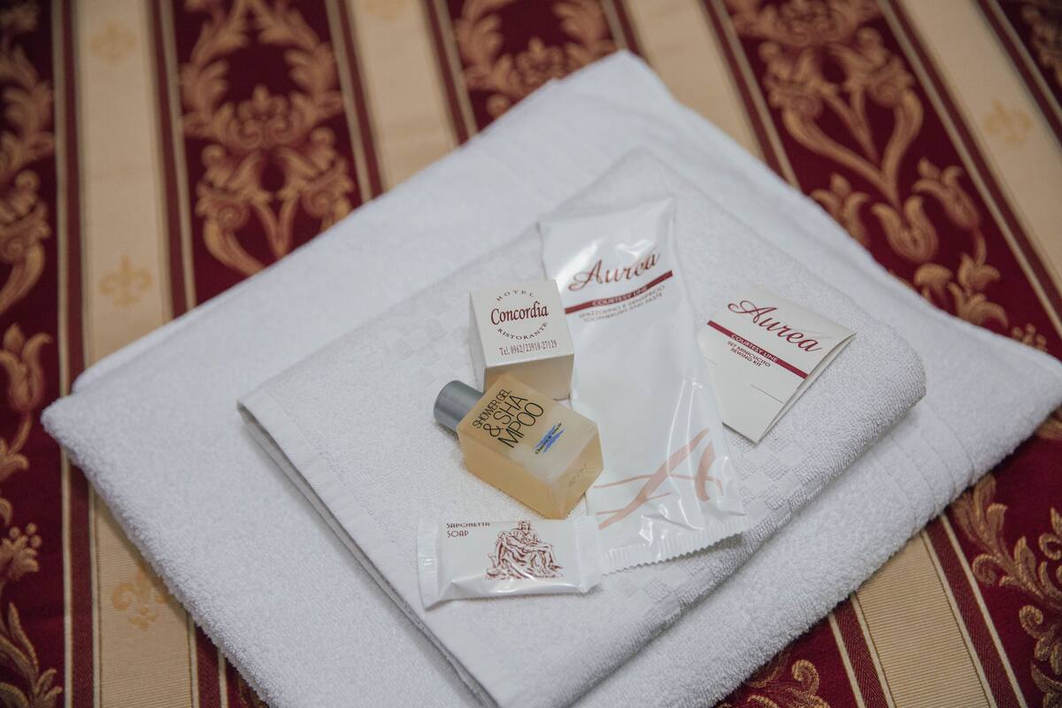 A neatly folded white towel with a bottle of shower gel, a bar of soap, and a sachet of body lotion on a patterned red and beige fabric.