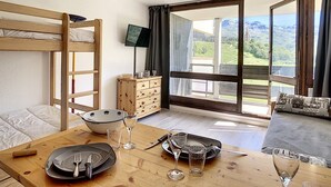 Una habitación con una cama individual superpuesta, una mesa de madera con cubiertos y vasos, una televisión y vistas a montañas a través de amplias ventanas.