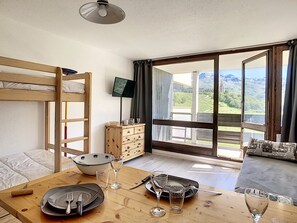 Een kamer met een stapelbed, een houten tafel met bestek en glazen, een televisie en uitzicht op bergen door grote ramen.