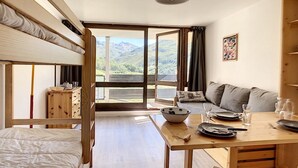 Una habitación acogedora con una cama, una mesa de madera con un bol y vasos, un sofá y vistas a montañas a través de amplios ventanales.