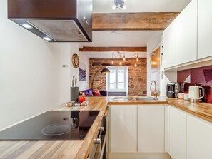 A modern kitchen with a central island, wooden beams, a stone wall, and a view to a living area. - Up-top Cottage (Holmfirth)