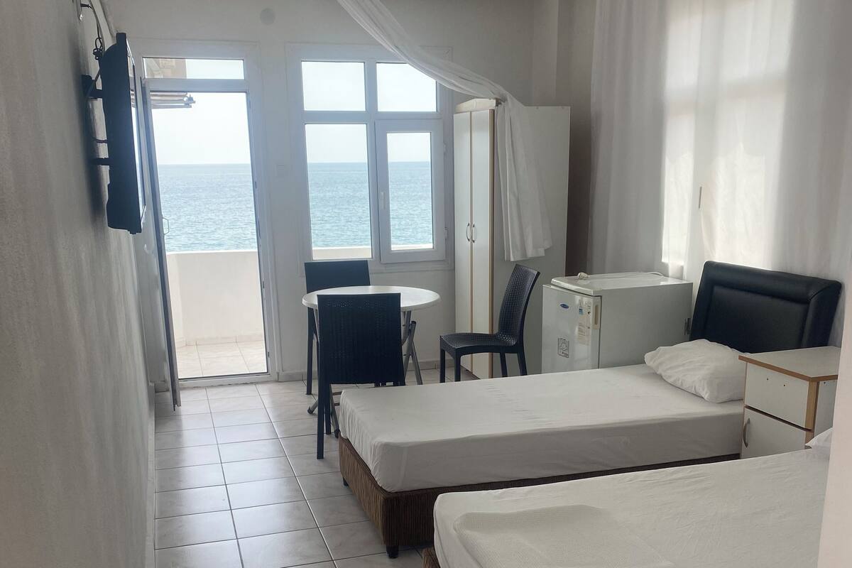 Quarto de hotel com duas camas, uma mesa pequena, uma cadeira, uma escrivaninha e vista para o mar.
