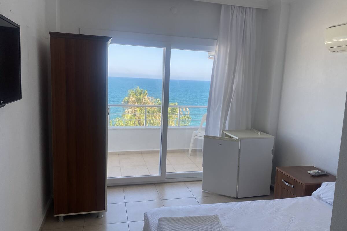 Quarto de hotel com cama, secretária pequena, frigorífico e porta de vidro deslizante para varanda com vista para o mar.