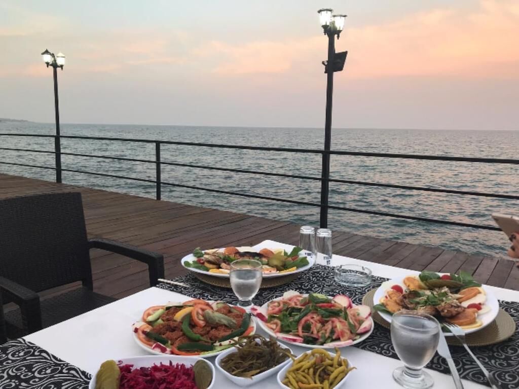 A table set with various dishes and drinks on a wooden deck overlooking the sea, with two street lamps in the background.