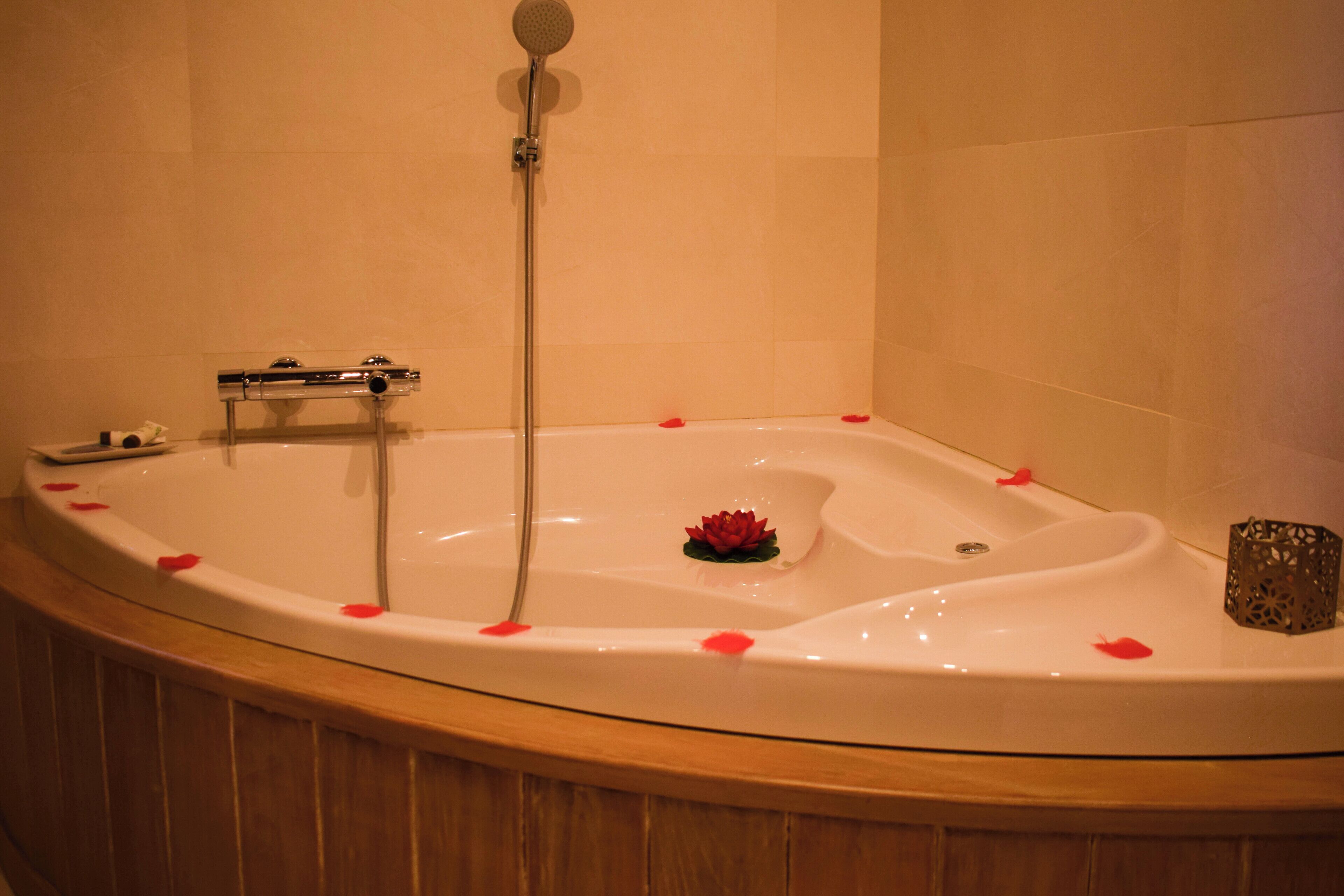 Una bañera con pétalos de rosa y una flor de loto, un cabezal de ducha manual y un candelabro.