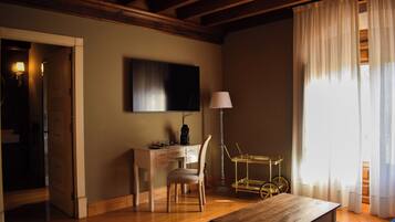 A room with a wooden desk, chair, and a television mounted on the wall. There is a lamp, a small table, and a window with curtains.